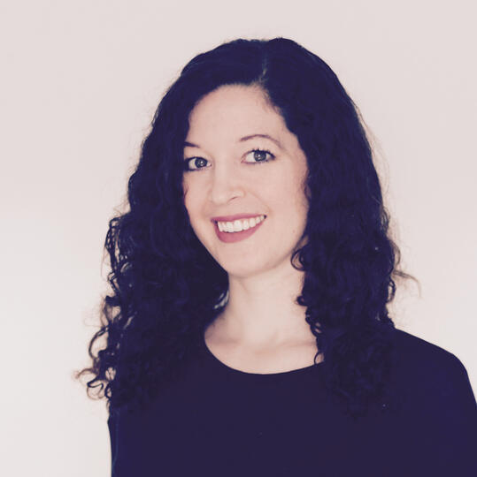 A portrait of a smiling white woman with dark, curly hair wearing a black shirt.